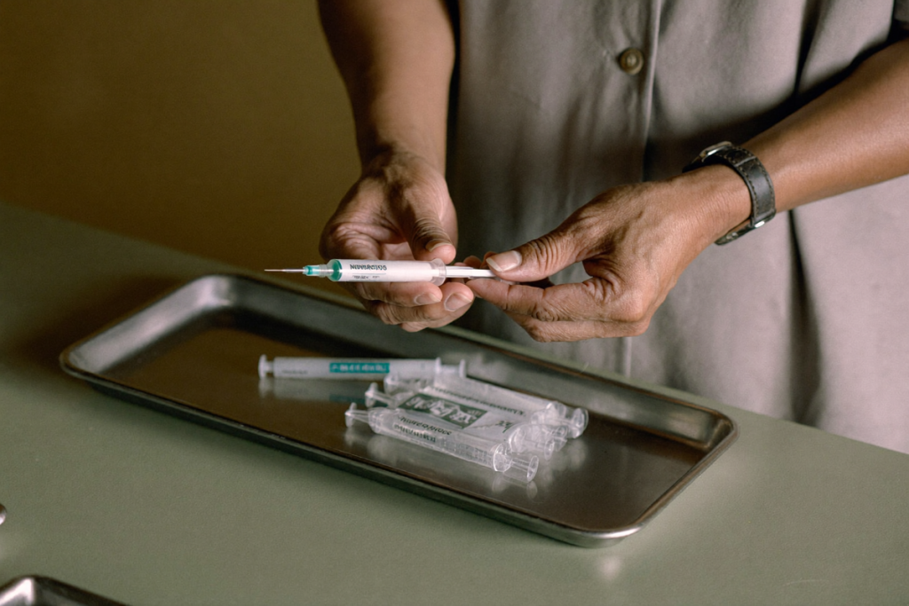 Gloved hands holding a sealed sterile syringe over a stainless tray on a sage-green counter