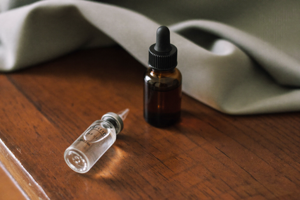 A clear glass vial resting beside an amber dropper bottle on a wooden surface, evoking an injectable route