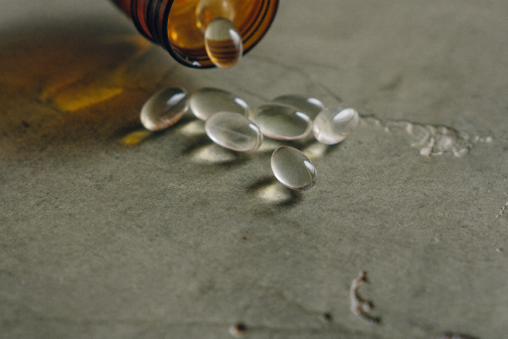 Translucent supplement capsules spilled from an amber bottle onto a sage-green surface, representing oral glutathione