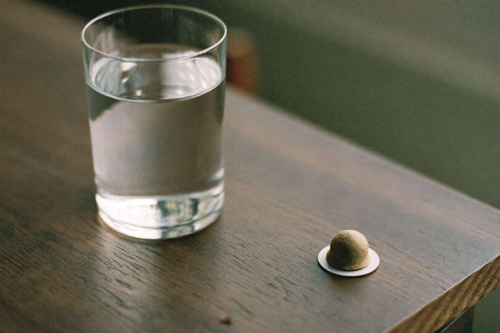 A glass of water and a single oral capsule on a wooden table in soft morning light