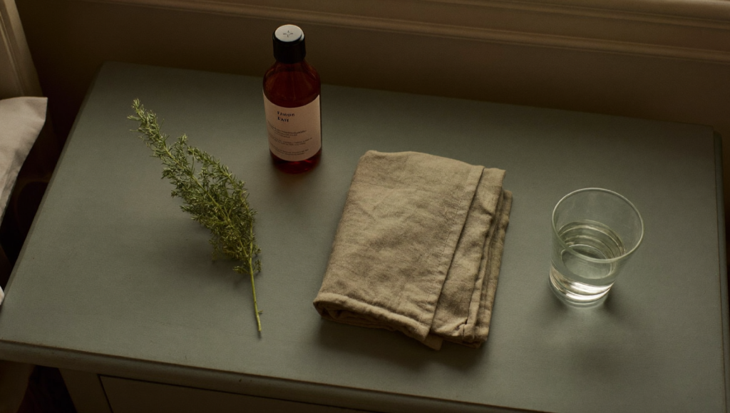 Calm bedside nightstand at dawn with a plain amber bottle, linen cloth, and a sprig of sage in sage-green and earth tones
