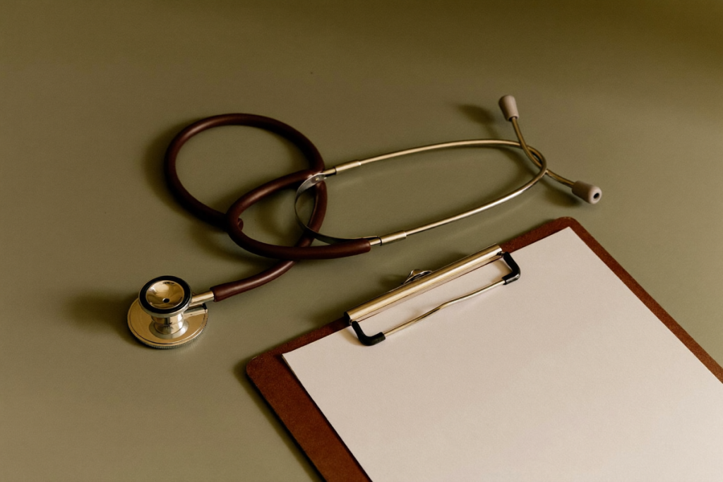 A stethoscope coiled beside a blank clipboard on a sage-green desk in soft daylight