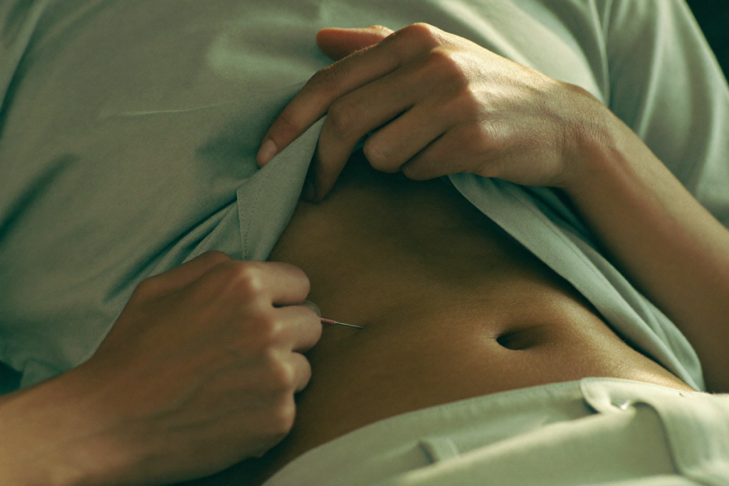 Hands pinching a fold of abdominal skin away from the navel to demonstrate a subcutaneous injection-site area