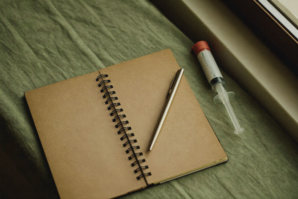 A blank notebook, pen, and a capped syringe on a sage-green cloth in window light