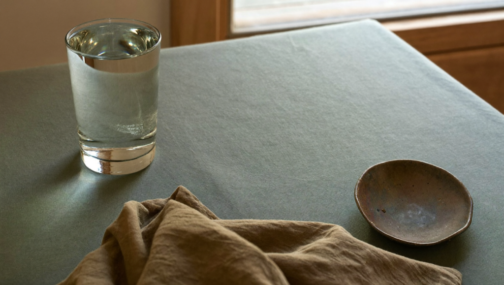 A glass of water and folded cloth on a sage-green surface in soft morning light, suggesting rest and hydration