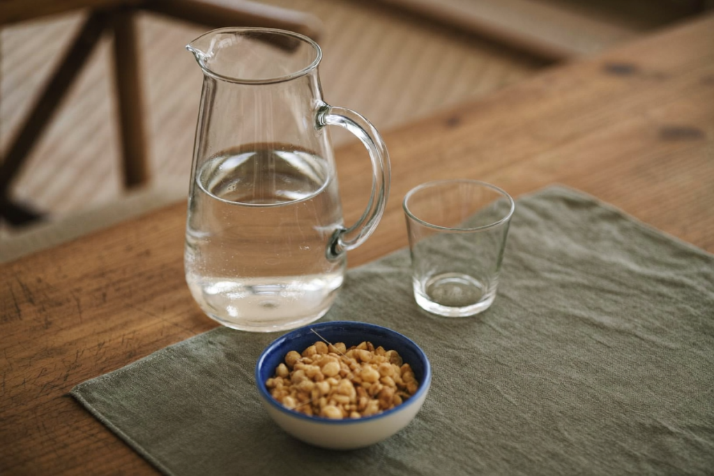 A water pitcher, glass, and small bowl of nuts on a wooden table in soft light
