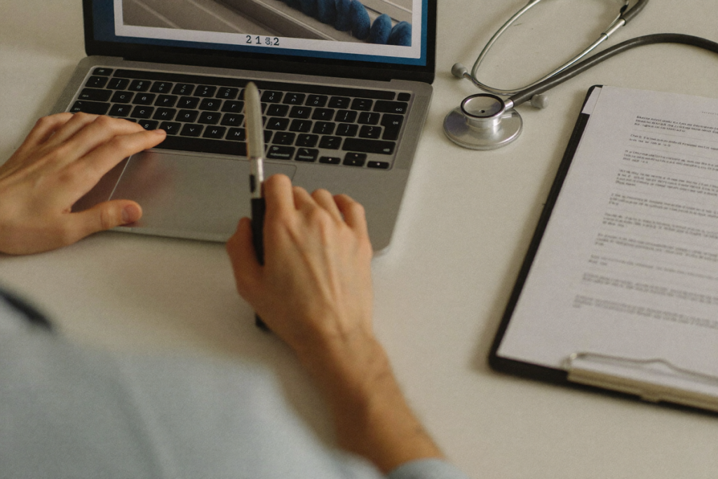 A clinician's hands at a laptop with a clipboard and stethoscope on a desk, representing a provider-led telehealth evaluation